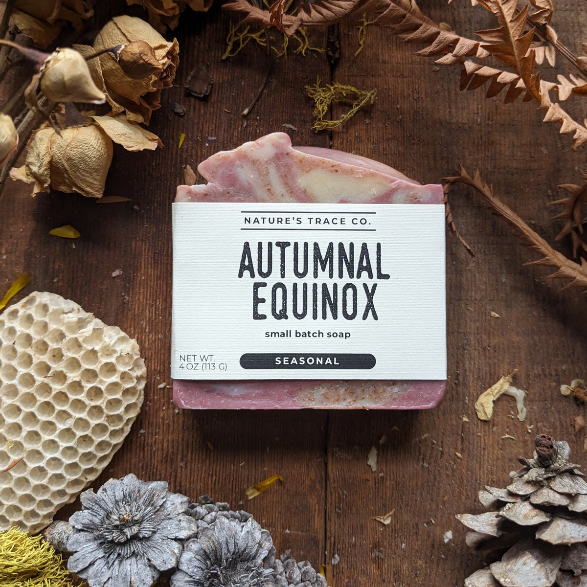 A bar of Nature's Trace Co. 'Autumnal Equinox' seasonal small batch soap, displayed on a rustic wooden background surrounded by dried botanicals, a honeycomb piece, and pinecones.